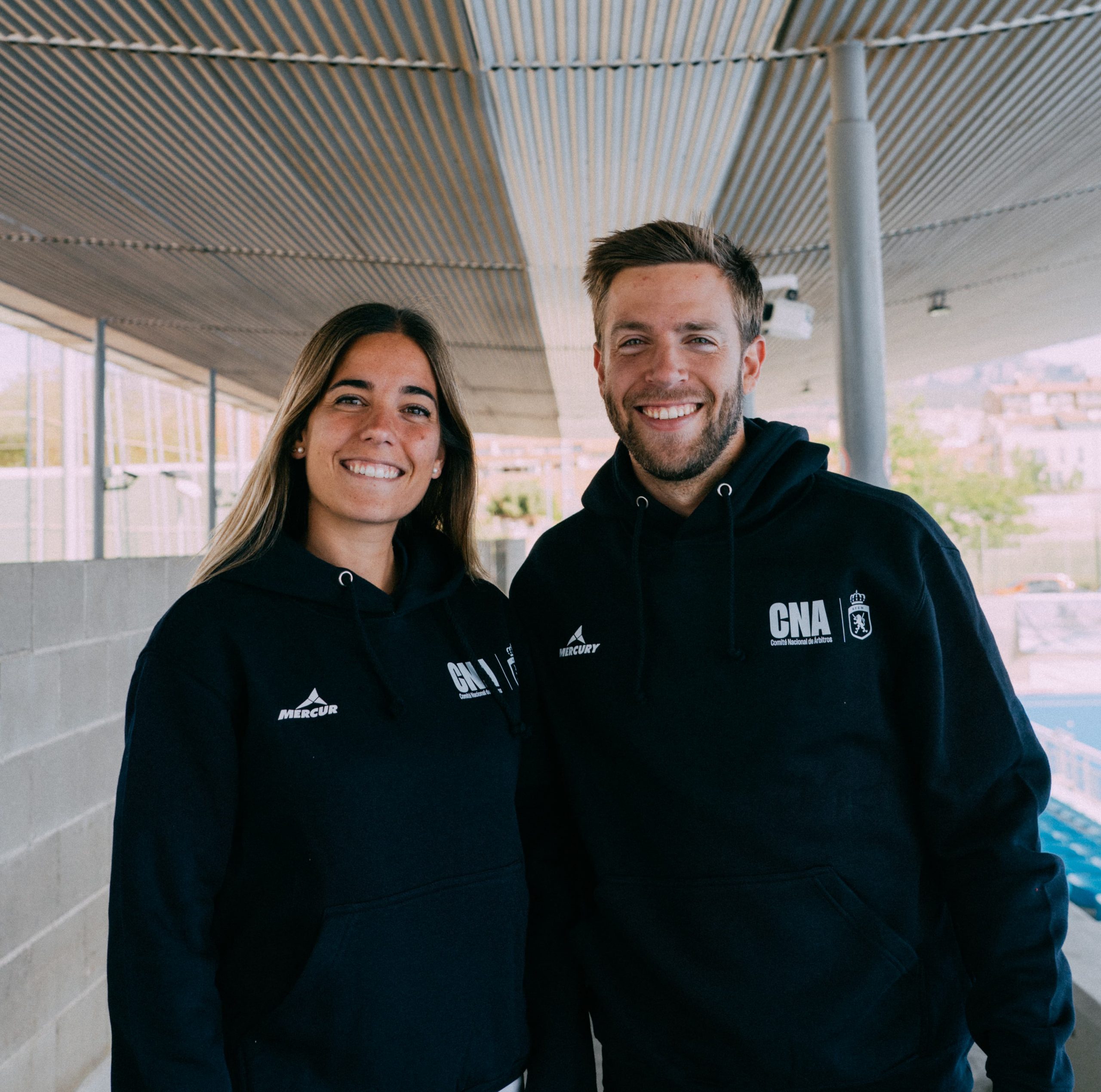 Chico y chica posan con ropa oficial de Mercury para árbitros de hockey hierba. Sudadera azul marino unisex del Comité Nacional de Árbitros
